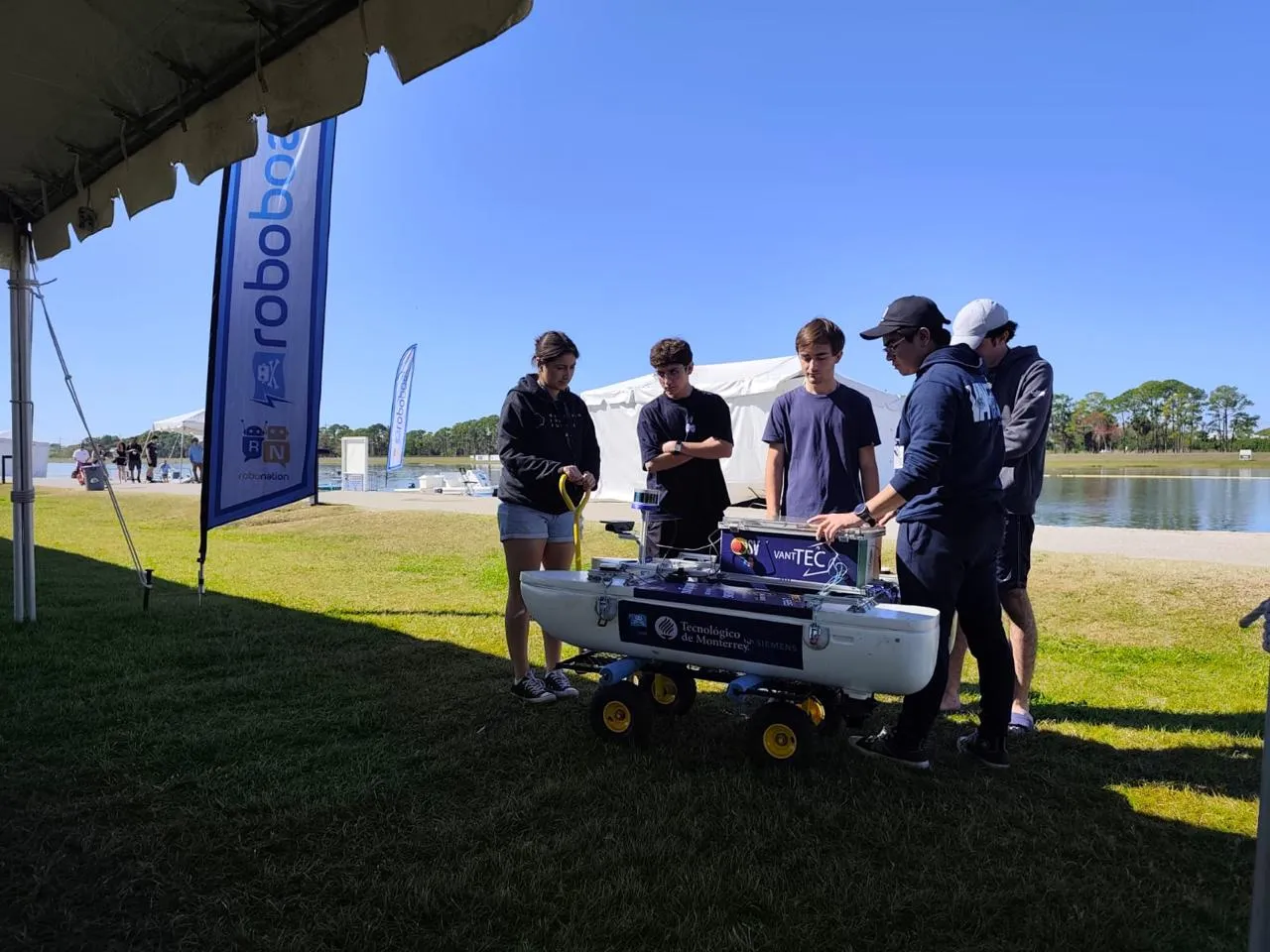 Five students standing around the white USV mounted on a wheeled cart on grass, with VANTTEC and Tecnológico de Monterrey branding on the hull and a RoboBoat feather flag beside them