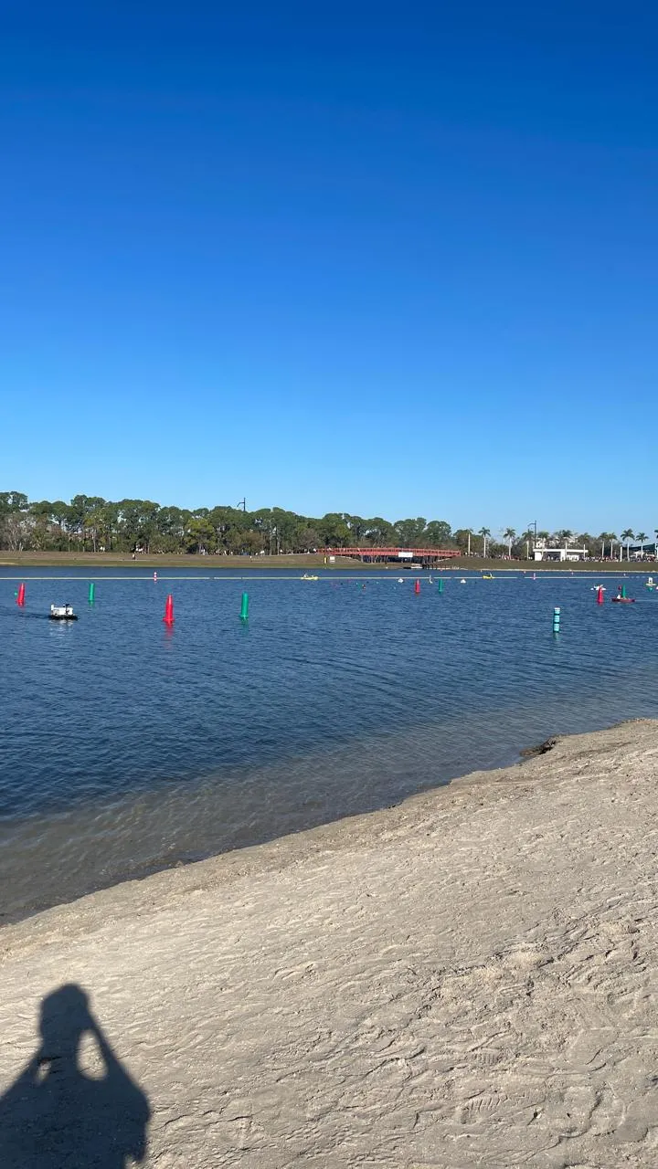 Wide view of a lake course with alternating red post buoys and green post buoys floating in lines on dark blue water, a small white robot boat at the left edge, and a red bridge spanning the far shore under a clear sky