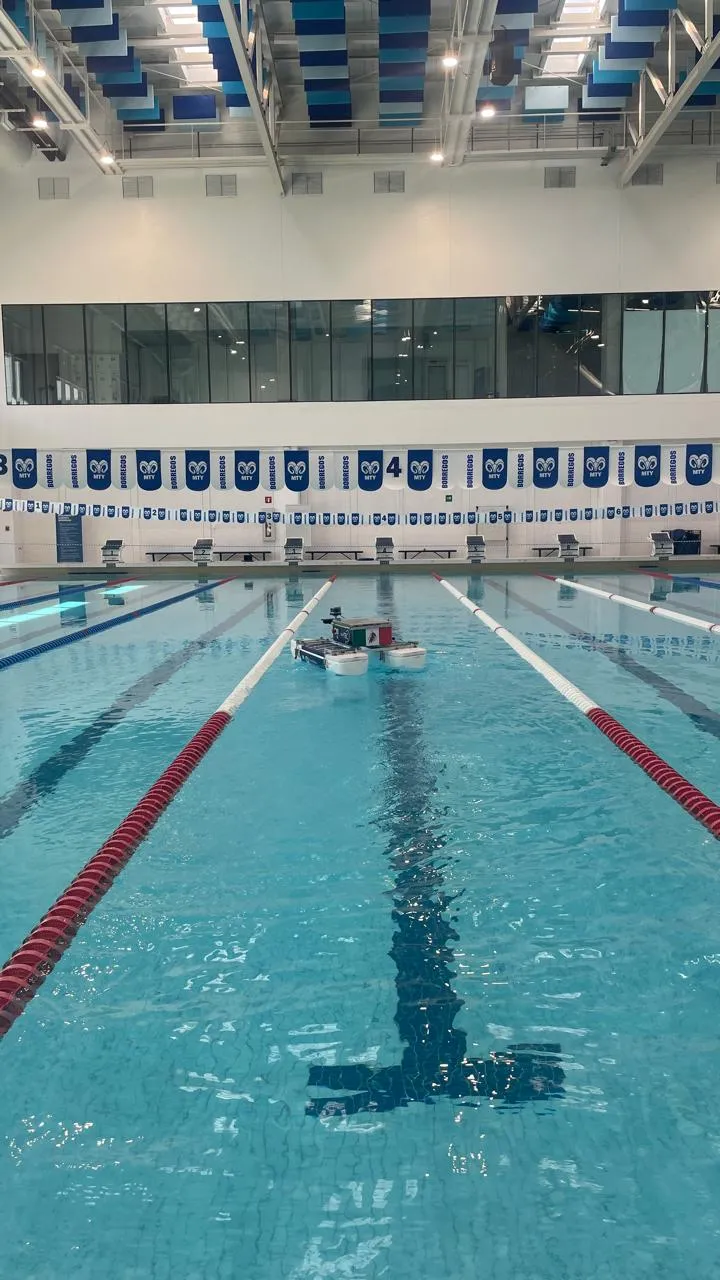 The white twin-hull robot boat floating in a lane of a large indoor swimming pool with Tec Borregos banners on the far wall