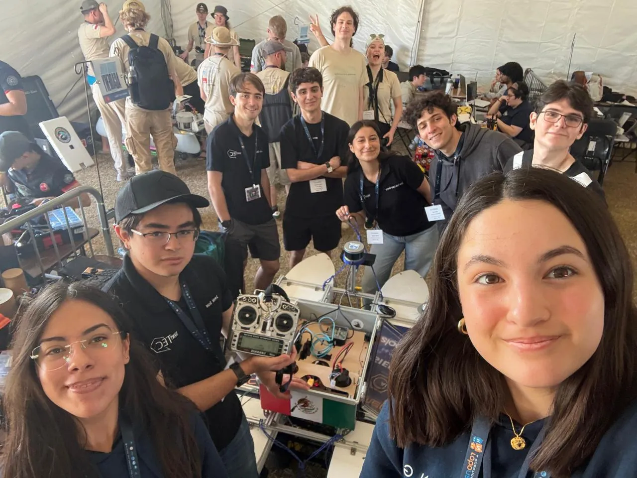 Group selfie of students in black team shirts and event lanyards gathered around a small white twin-hull robot boat on a stand, one student holding an RC transmitter, with wiring and electronics visible inside the open hull and a work tent behind them